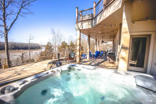A hot tub on a deck overlooking a river, with outdoor seating and trees in the background under a clear blue sky.