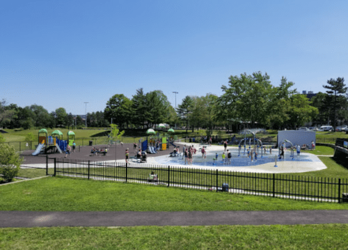 A sunny park scene featuring a playground and a splash pad, with children playing and families enjoying the outdoors.
