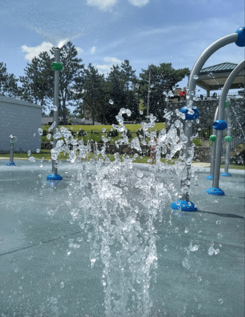 A splash of water sprays up from a fountain in a sunny playground, surrounded by greenery and play structures.