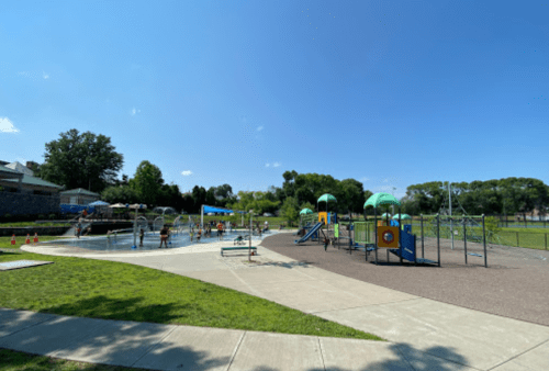 A sunny park featuring a splash pad and playground with children playing and green trees in the background.