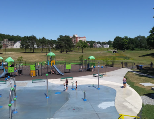 A sunny playground with green equipment, children playing, and a grassy area in the background.