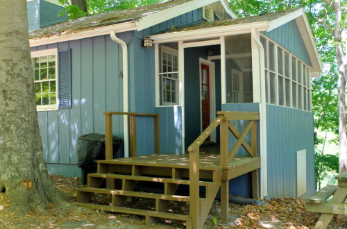 A small blue cottage with a screened porch and wooden steps, surrounded by trees and greenery.