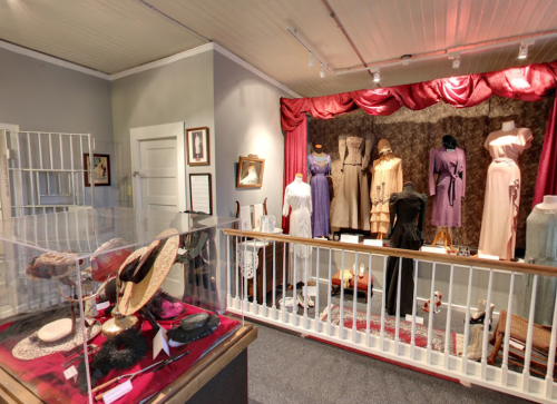 A display of vintage dresses and accessories in a museum setting, featuring mannequins and a glass case.