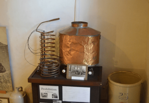 Copper distilling apparatus displayed on a wooden stand, with a historical photo and prohibition-related documents nearby.