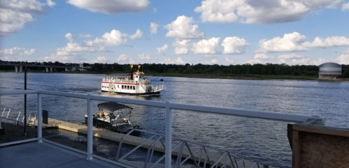 A riverboat sails on a calm river under a blue sky with fluffy clouds, with a dock and boat in the foreground.