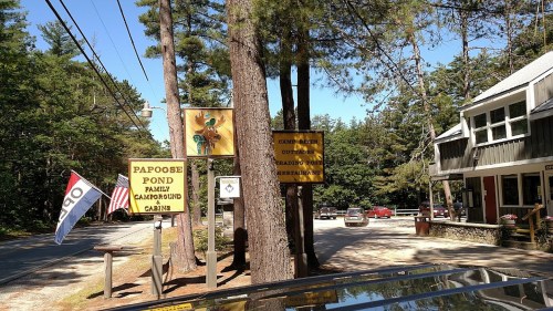 Signage for Papoose Pond Family Campground, with trees and a building in the background, and an American flag visible.