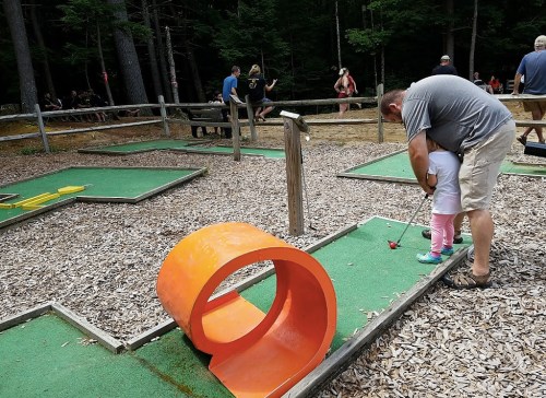 A man helps a child at a mini-golf course, with colorful obstacles and trees in the background.