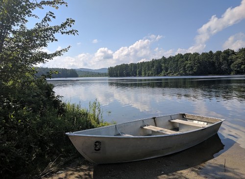 A small boat rests on the shore of a calm lake, surrounded by trees and a clear blue sky with fluffy clouds.