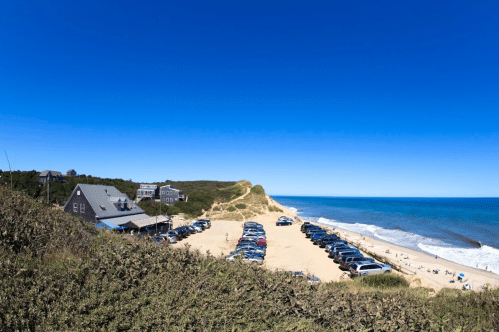 A panoramic view of a beach with parked cars, dunes, and a clear blue sky.