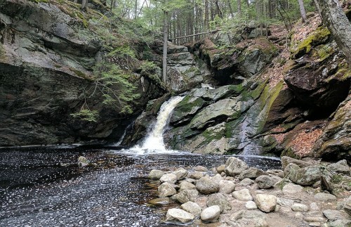 A serene waterfall cascades over rocks into a dark pool, surrounded by lush greenery and rugged cliffs.