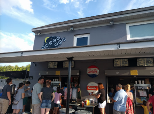 A busy snack bar with a line of customers waiting to order, under a blue sky with scattered clouds.