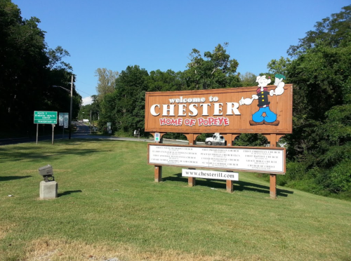 Sign welcoming visitors to Chester, home of Popeye, with green directional signs and trees in the background.
