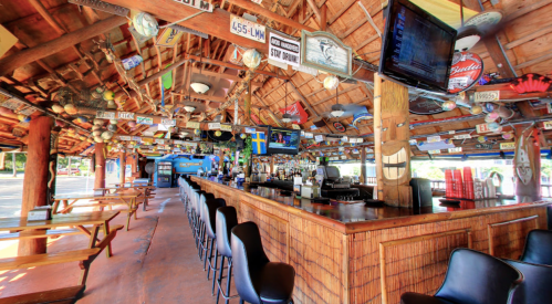 A vibrant bar interior with wooden decor, signs, and a long counter, surrounded by tables and a tropical atmosphere.