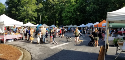 A bustling outdoor market with people browsing stalls, tents, and dogs, surrounded by trees on a sunny day.