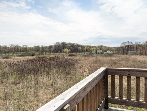 A wooden railing overlooks a vast, open field with sparse vegetation and distant trees under a cloudy sky.