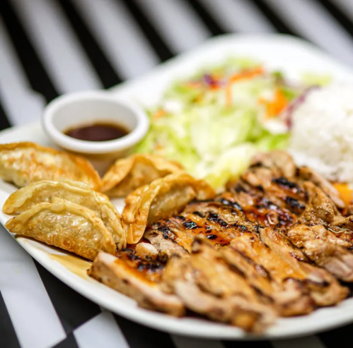 A plate of grilled chicken, dumplings, salad, and rice, served with a small bowl of dipping sauce.