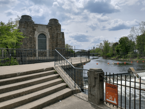 A stone building by a river with a staircase, railing, and a warning sign about water safety. Cloudy sky above.