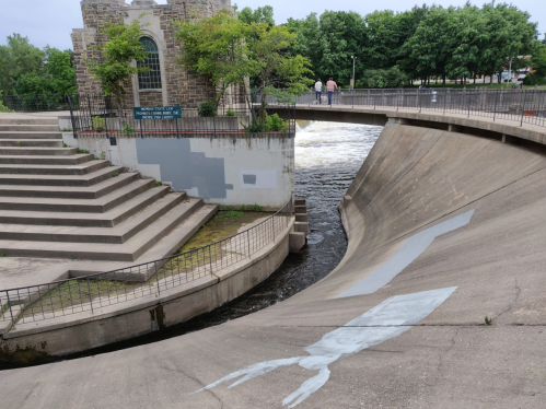 A concrete channel with a painted design, leading to a river, surrounded by greenery and steps. People walk in the background.