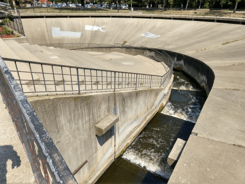A concrete channel with flowing water, surrounded by sloped banks and a railing, under a clear blue sky.