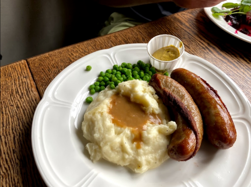 A plate with mashed potatoes, green peas, two sausages, and a small cup of mustard on a wooden table.