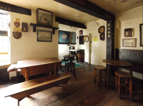 A cozy pub interior featuring wooden tables, stools, and dartboard, with vintage decor on the walls.