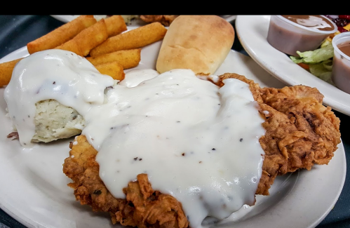 A plate of fried chicken topped with creamy gravy, served with a biscuit, fries, and sides of salad and sauce.