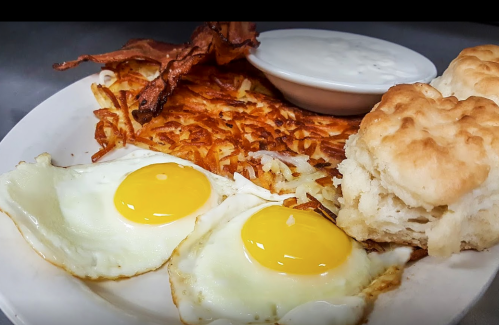 A plate of breakfast featuring two sunny-side-up eggs, crispy bacon, hash browns, a biscuit, and a small bowl of gravy.