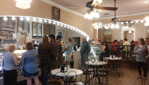 A busy café interior with customers at the counter, tables, and a display of desserts under warm lighting.