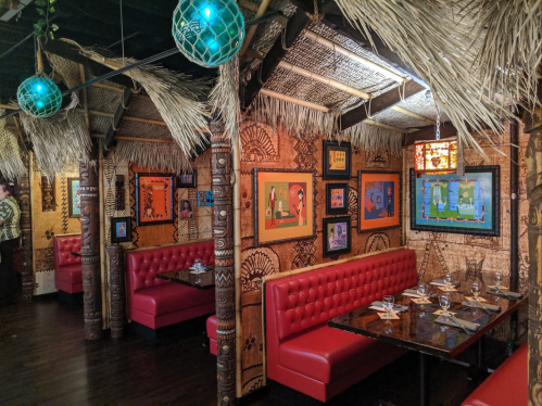 Tropical-themed restaurant interior with red booths, thatched roof, and colorful artwork on the walls.