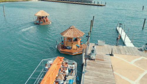 Two boats, one with a thatched roof, navigate a calm blue waterway near a wooden dock.