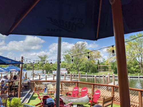 A riverside patio with umbrellas, chairs, and string lights, overlooking a calm waterway and greenery.