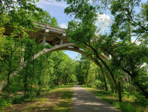 A concrete bridge arches over a tree-lined path, surrounded by lush greenery and blue skies.