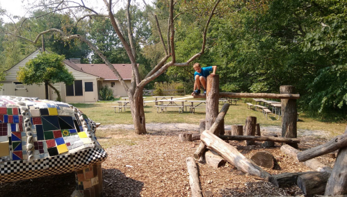 A child sits on a wooden post in a playground surrounded by trees and picnic tables.
