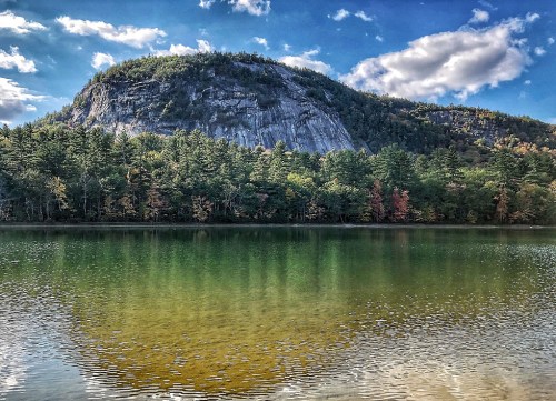 A serene lake reflects a rocky mountain and colorful trees under a blue sky with fluffy clouds.