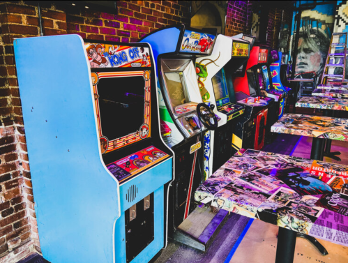 A row of vintage arcade machines in a brick-walled venue, with colorful cabinets and tables covered in comic book pages.
