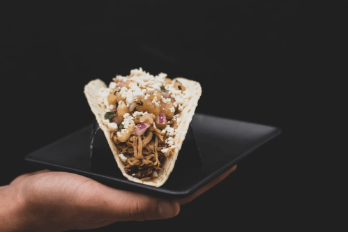 A hand holding a taco filled with shredded meat, topped with diced onions and cheese, on a black plate.