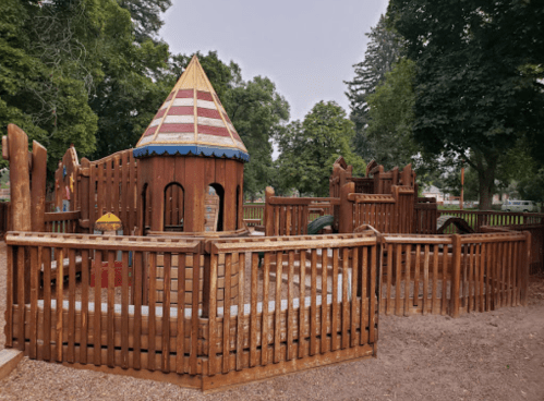 A wooden playground with a castle-like structure, surrounded by a fence and trees in the background.