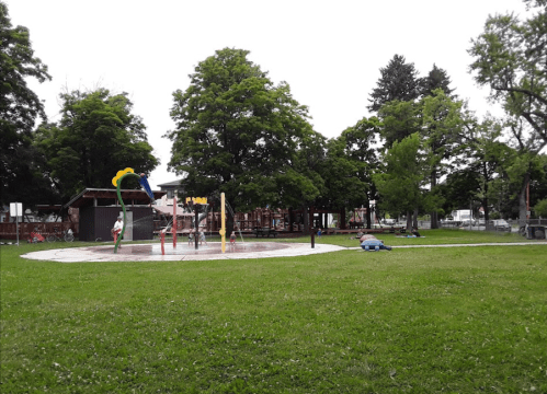 A park scene featuring a splash pad surrounded by trees and grassy areas, with a few people enjoying the space.