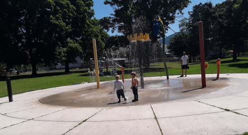 Two children play in a splash pad at a park, with water spraying around them and a person standing nearby.