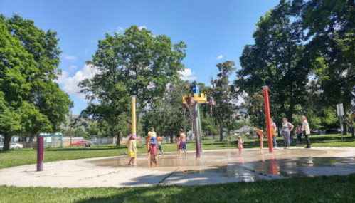 Children play in a splash pad at a park on a sunny day, surrounded by trees and adults watching nearby.