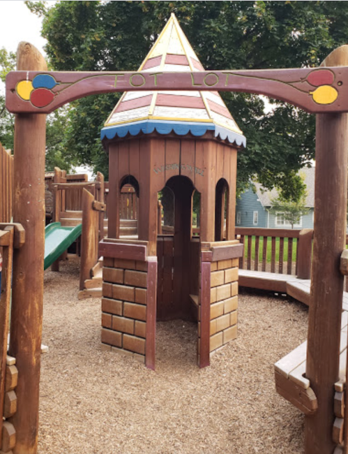 A wooden playhouse with a peaked roof and colorful accents, surrounded by playground equipment and gravel.