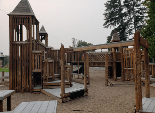 A wooden playground structure with towers, slides, and climbing features, set in a gravel area surrounded by trees.