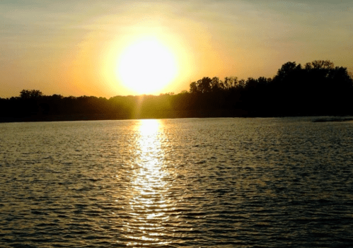 Sunset over a calm lake, with golden light reflecting on the water and silhouettes of trees in the background.
