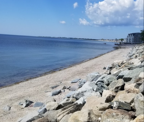 A serene beach scene with calm water, rocky shoreline, and a clear blue sky dotted with clouds.