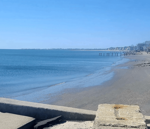 A serene beach scene with calm blue waters, sandy shore, and distant buildings under a clear blue sky.