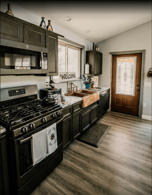 A modern kitchen with dark cabinets, a farmhouse sink, and a welcoming entrance door. Natural light fills the space.