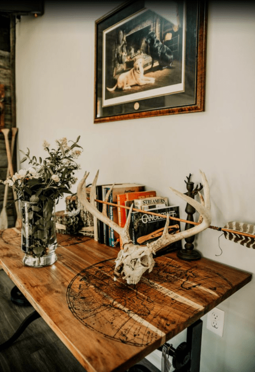 A rustic wooden table with a deer skull, flowers in a vase, books, and a framed artwork on the wall.