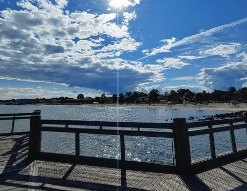 A sunny beach scene with a wooden pier, sparkling water, and scattered clouds in a blue sky.