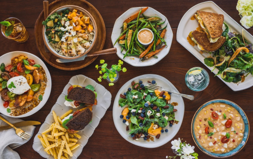 A variety of colorful dishes including salads, sandwiches, soups, and sides arranged on a wooden table.