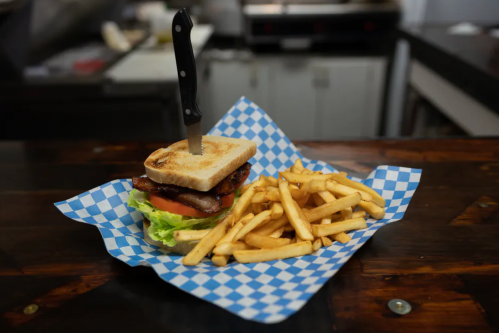 A sandwich with a knife in it, served on a blue checkered paper, alongside a pile of golden French fries.